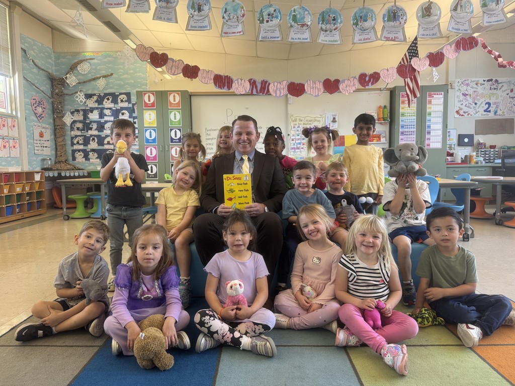 A group of young children and an adult seated on a classroom floor, surrounded by colorful decorations and educational posters.