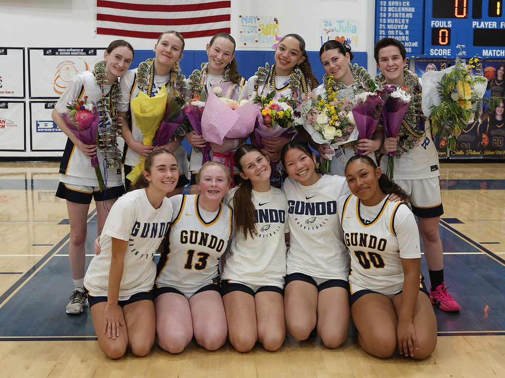High school girls basketball team posing on court with bouquets and leis after a game, wearing Gundo jerseys.