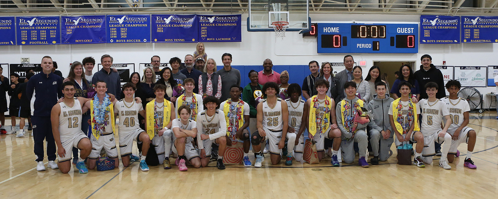 High school basketball team posing with coaches and supporters on gym court after a game, all smiling for a group photo.