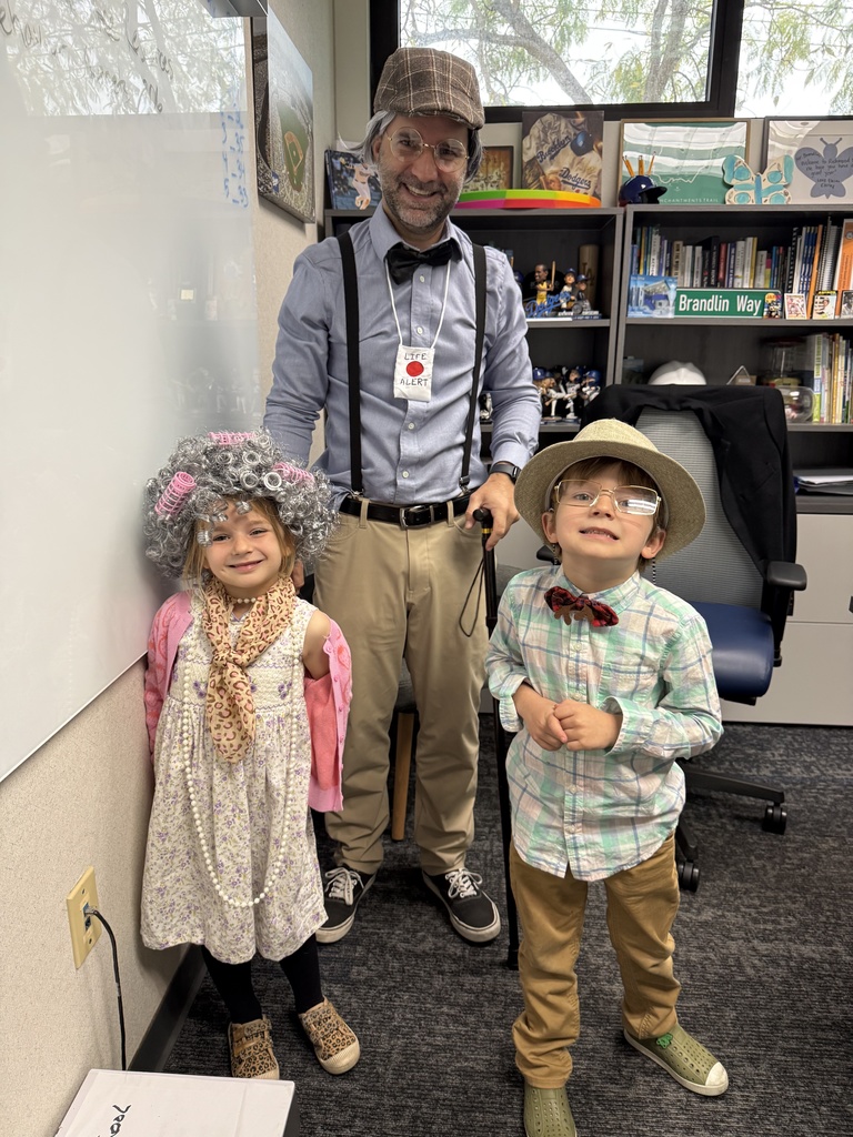 Two children and a teacher dressed in vintage-style costumes standing in an office with bookshelves and a whiteboard behind them.
