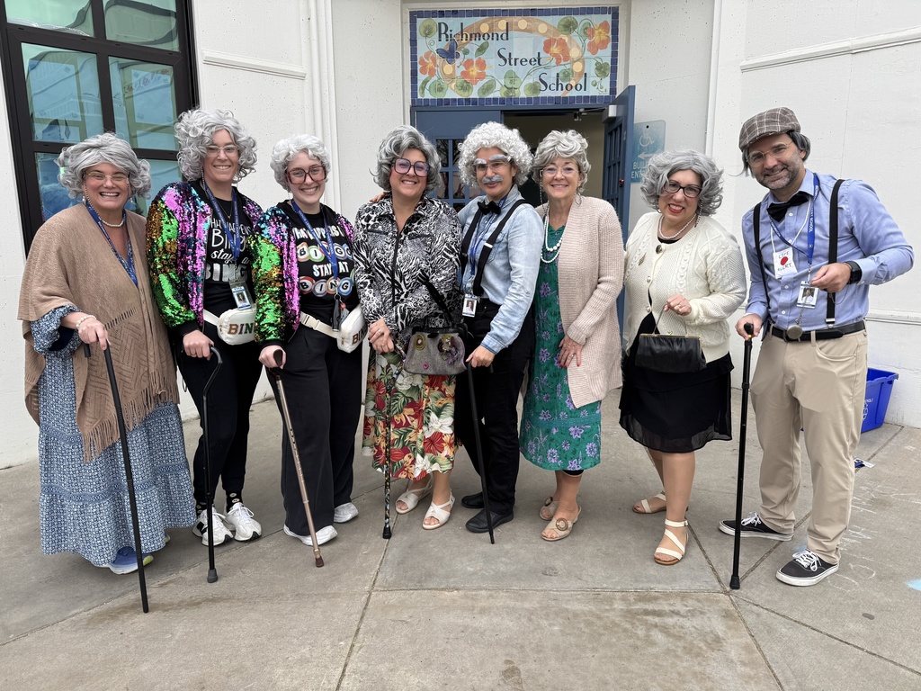 Group of teachers dressed as elderly individuals standing outside Raymond Street School, dressed in colorful and patterned clothing, some using canes for support.