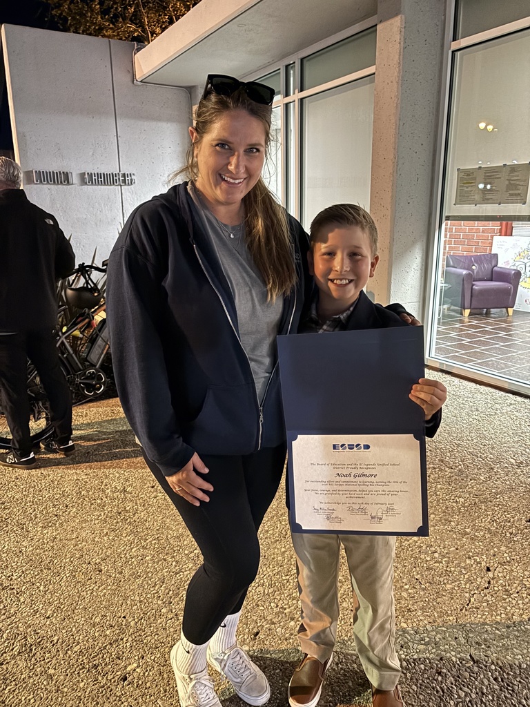 Woman and smiling boy stand outside at night, boy holding framed school award certificate.