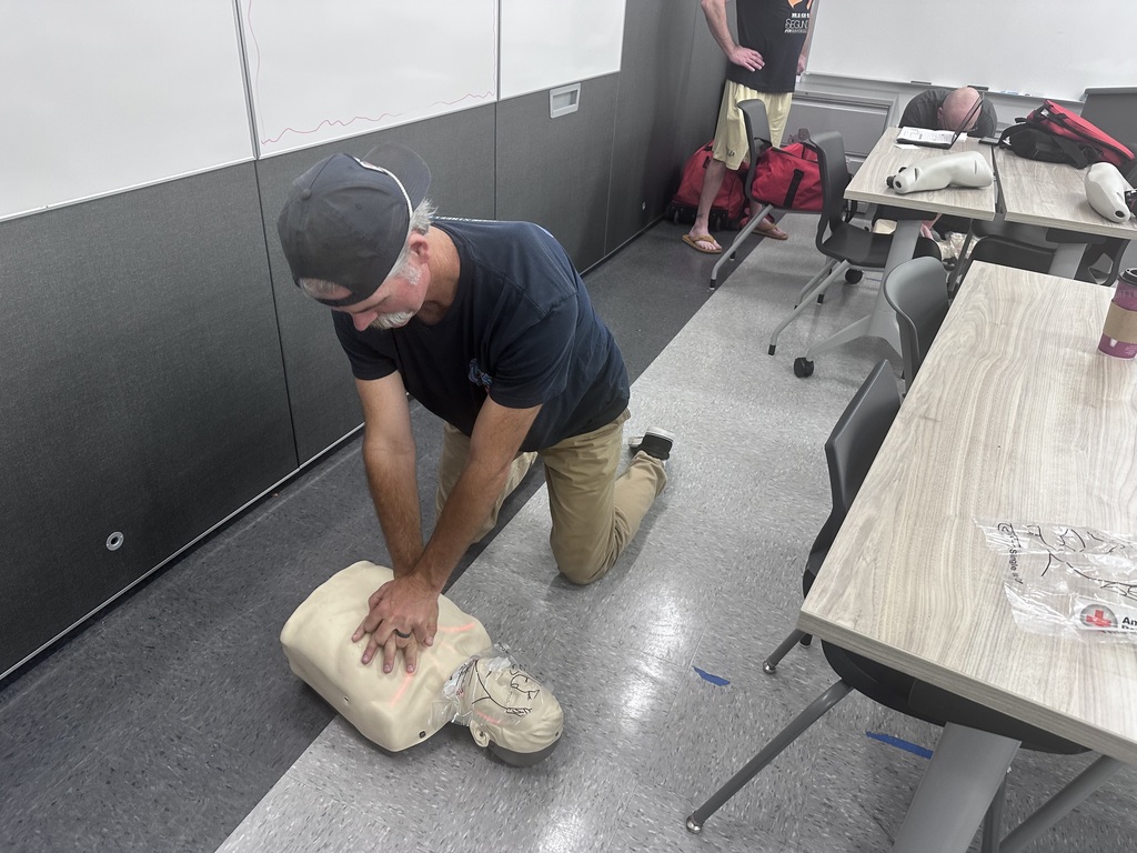 Man in cap kneels on classroom floor performing chest compressions on a CPR training mannequin.