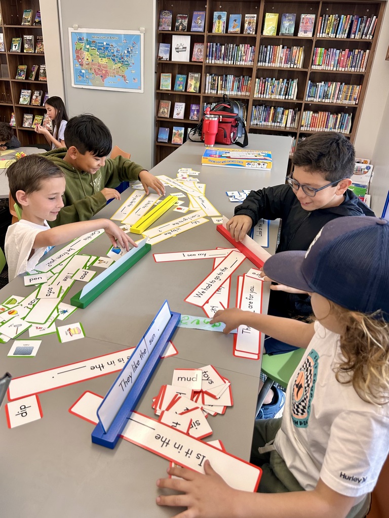 Students sit around a table in a library, working together to build sentences using word cards and learning materials.
