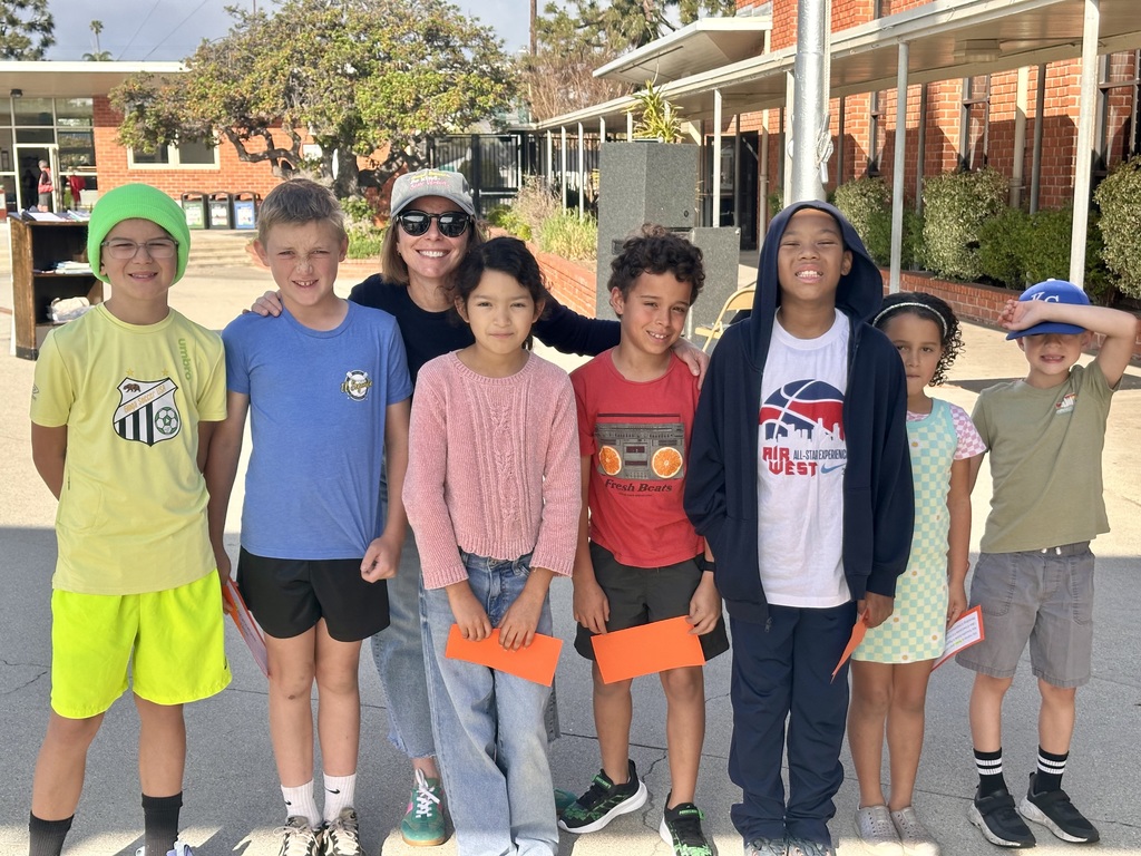 A teacher stands with a group of smiling elementary students outdoors, posing together on a sunny school campus.