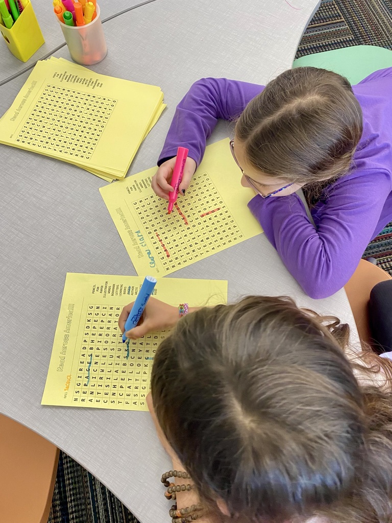 Two students use bright markers to complete word search worksheets at a classroom table with stacks of papers nearby.