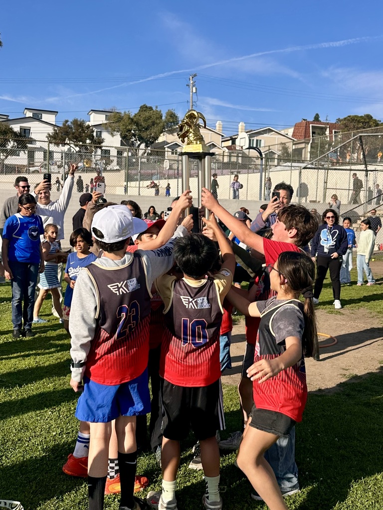 Students gather together on a field, raising a trophy in celebration while others watch and take photos nearby.