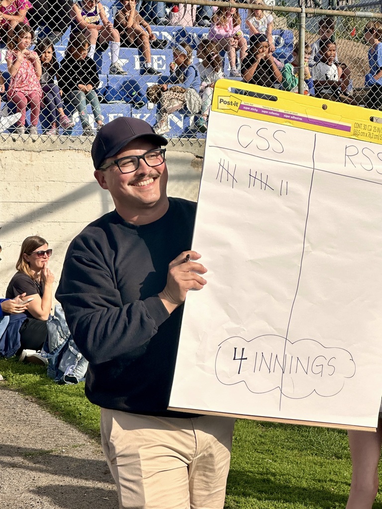 A staff member holds a score sheet showing innings and tallies while students sit and watch from bleachers behind.