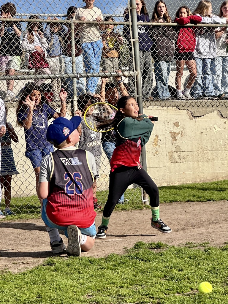 A student follows through on a swing as a ball drops nearby, with teammates and spectators watching closely.