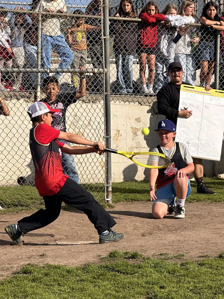 A student swings at a ball during a game while a teammate kneels as catcher and classmates watch from behind a fence.