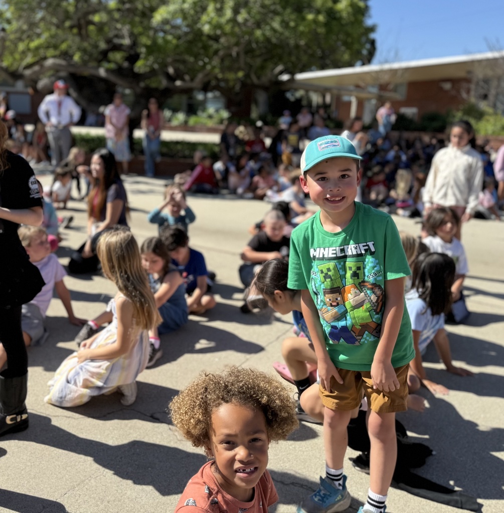 Two young students smile in the foreground while classmates sit on the ground during a sunny outdoor school assembly.