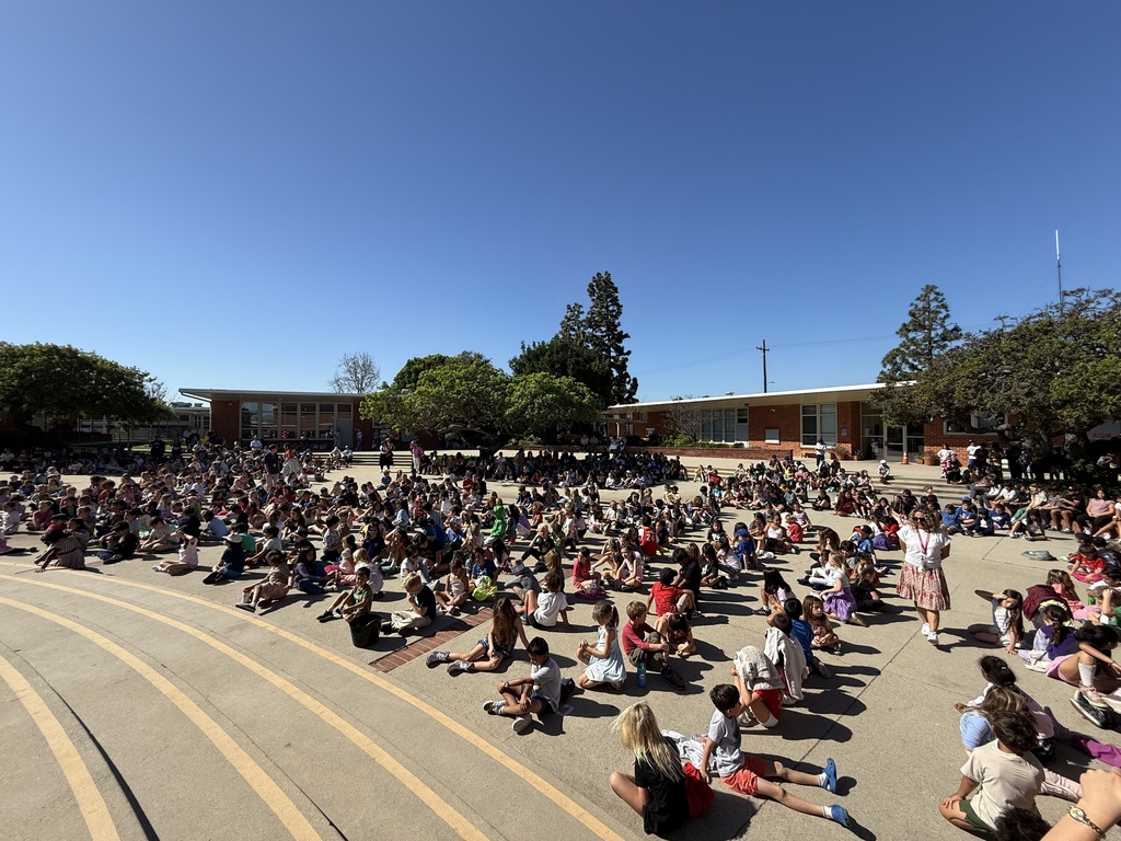 A large group of students sit spread out across a sunny courtyard, watching a school assembly.
