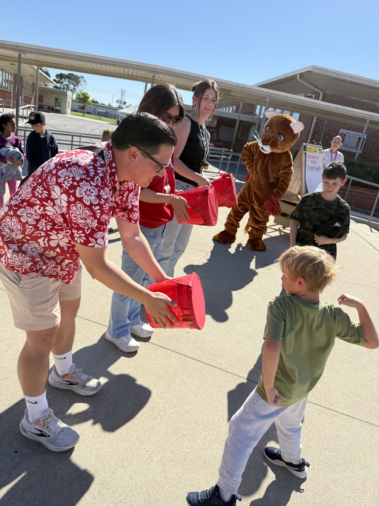 Staff hand out red-wrapped prizes to students during an outdoor event as a mascot stands nearby cheering.