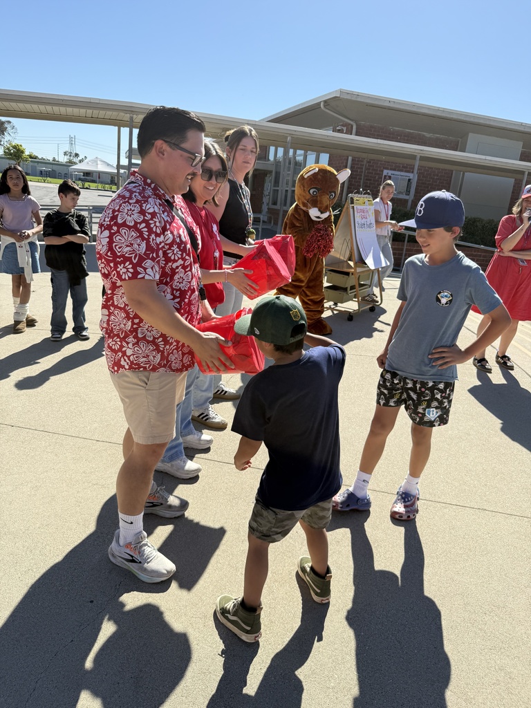 A student receives a red-wrapped prize from staff while others wait nearby, with a school mascot and announcer in the background.