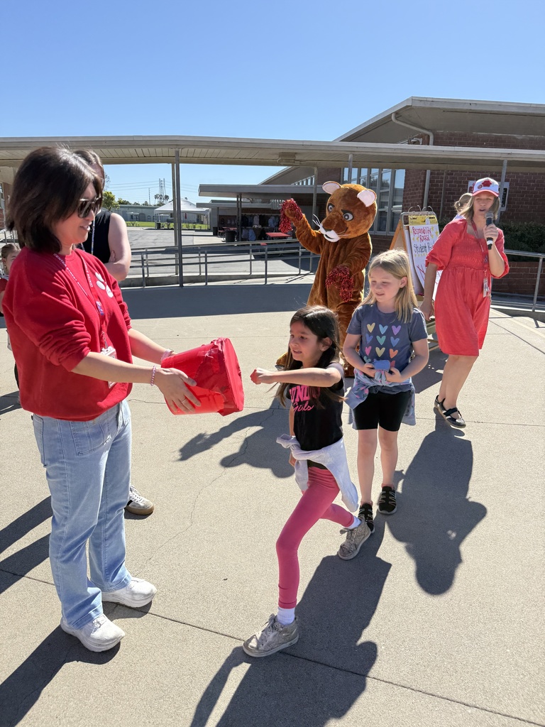Staff hand out red-wrapped prizes to students during an outdoor event as a mascot stands nearby cheering.