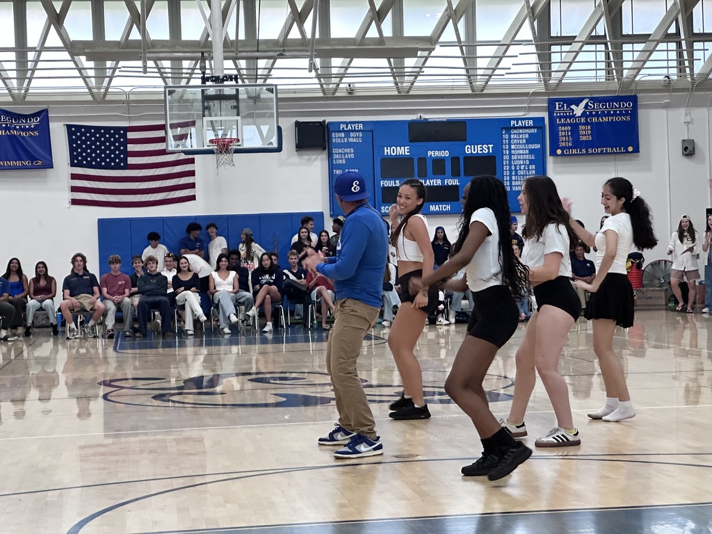 Students and a staff member dance together on a gym floor while classmates watch from bleachers beneath banners and an American flag.