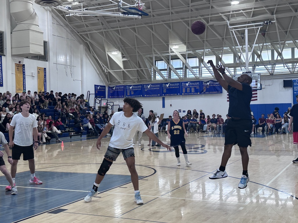 A player jumps to shoot a basketball during a gym activity as classmates and spectators watch from the sidelines and bleachers.