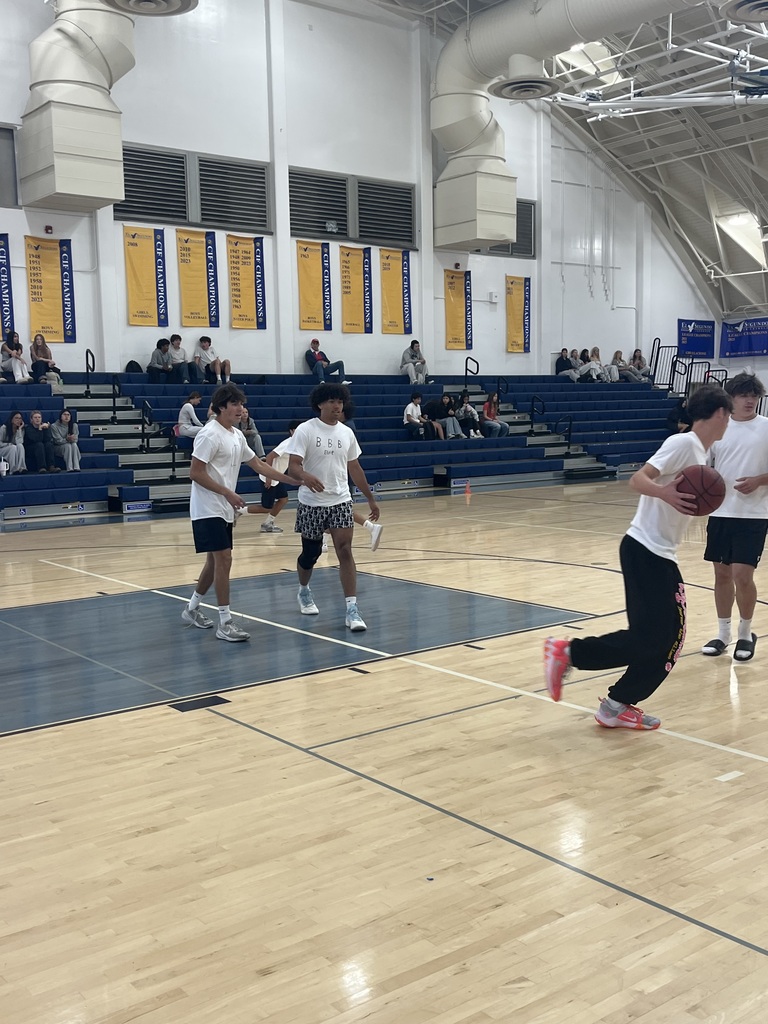 Students play basketball in a school gym, with others sitting on blue bleachers and championship banners lining the walls.