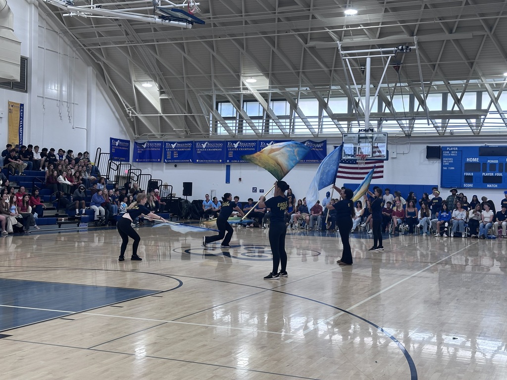 Students perform a color guard routine in a gym, waving large blue and gold flags while an audience watches.
