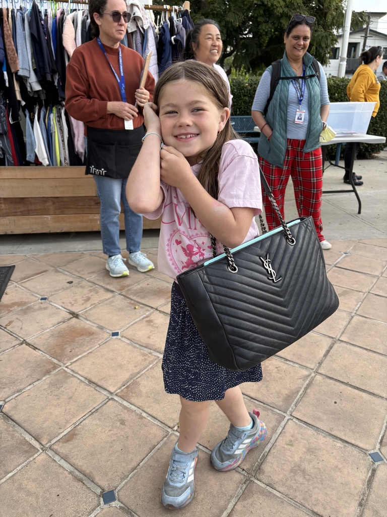 Young girl posing outdoors with a large black YSL handbag over her shoulder, wearing a pink shirt and sneakers.