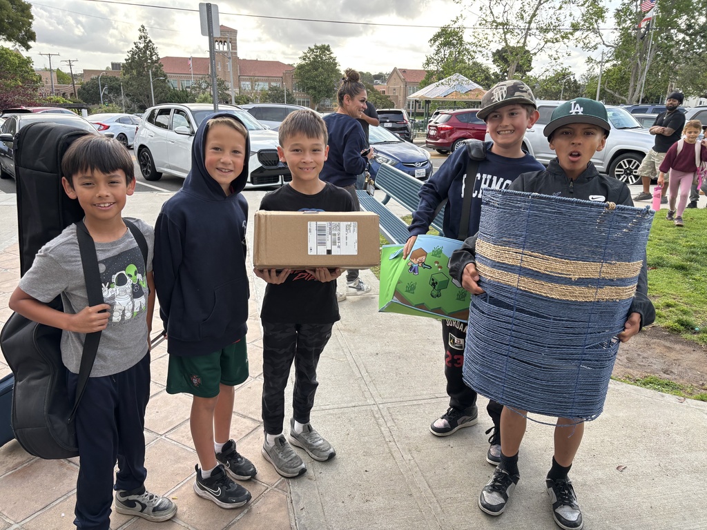 Group of boys standing outdoors, one holding a cardboard box, another carrying a rolled-up mat, and others with backpacks and bags.