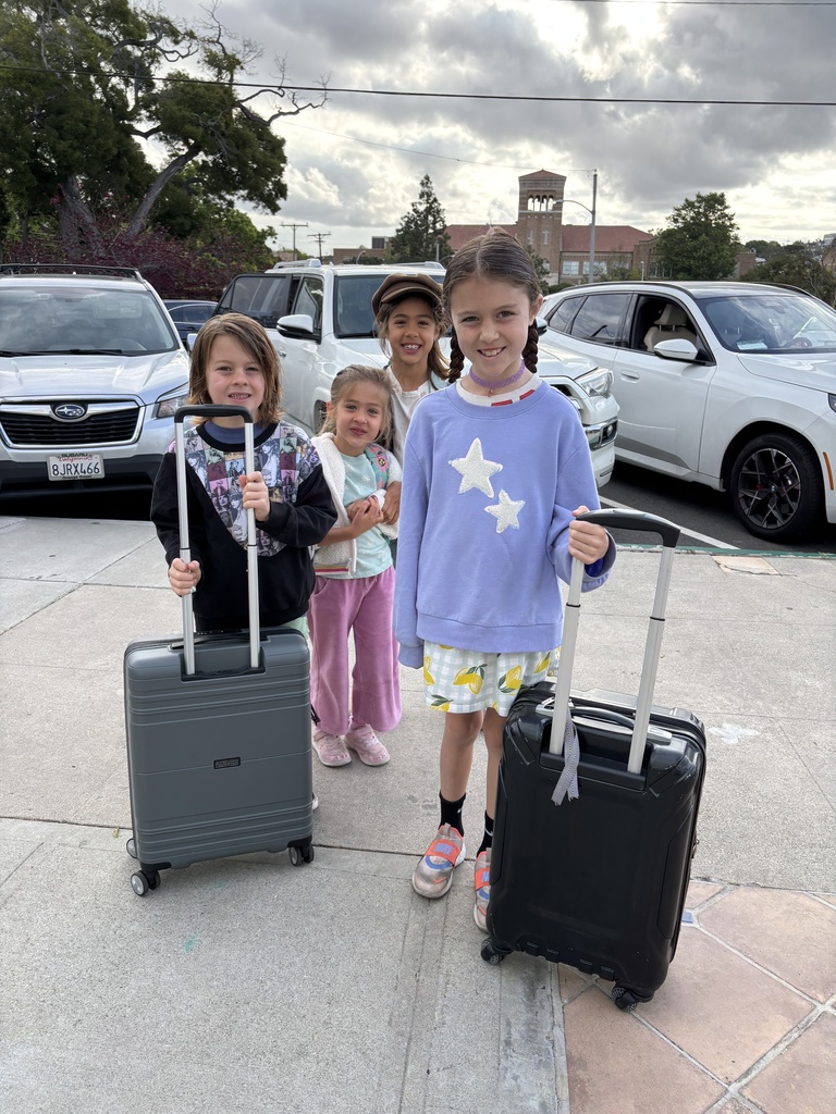 Four children standing on a sidewalk with rolling suitcases, ready for travel on a cloudy day.