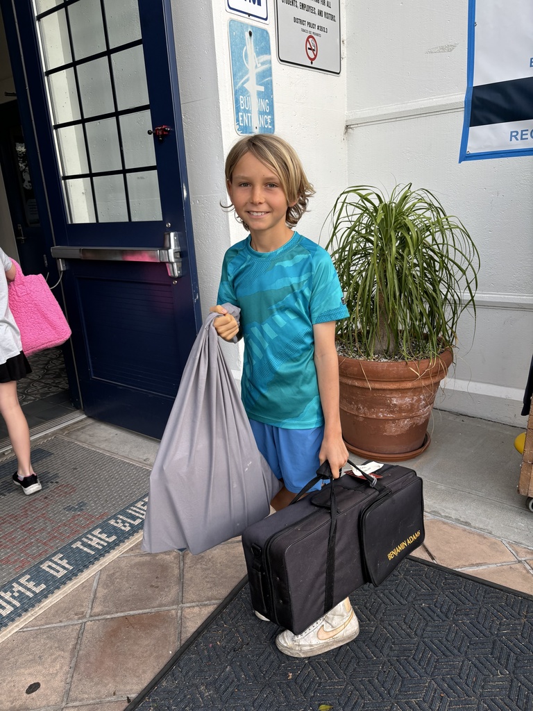 Young boy in blue shirt and shorts carrying a large gray bag and a black case outside a building entrance.
