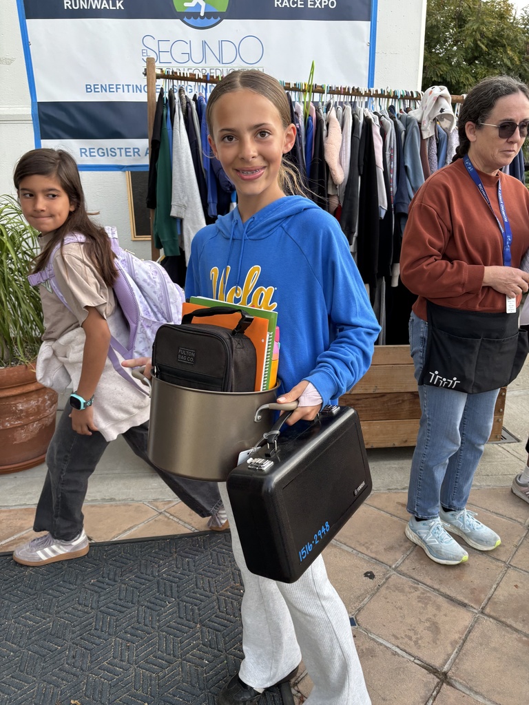 Student in a blue UCLA hoodie holding a cooking pot filled with books and a black case, standing outside near a clothing rack at an event.