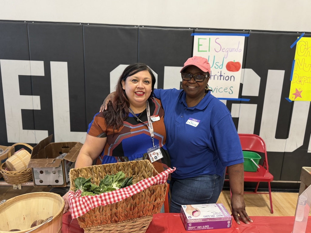 Two women smile with arms around each other behind a table displaying fresh leafy greens in a gym setting.