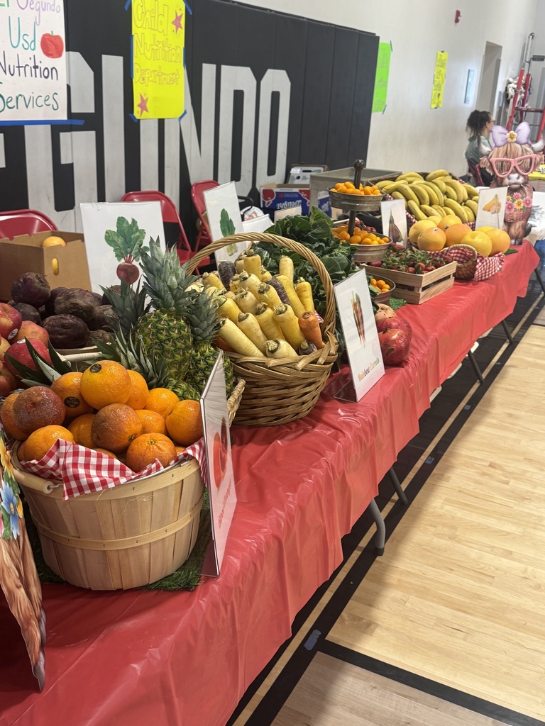 Long table covered in baskets of colorful produce including oranges, bananas, carrots, and beets, arranged for display.