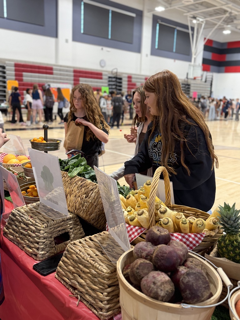 Students browse baskets of fruits and vegetables at a table in a gym, selecting items and placing them into bags.