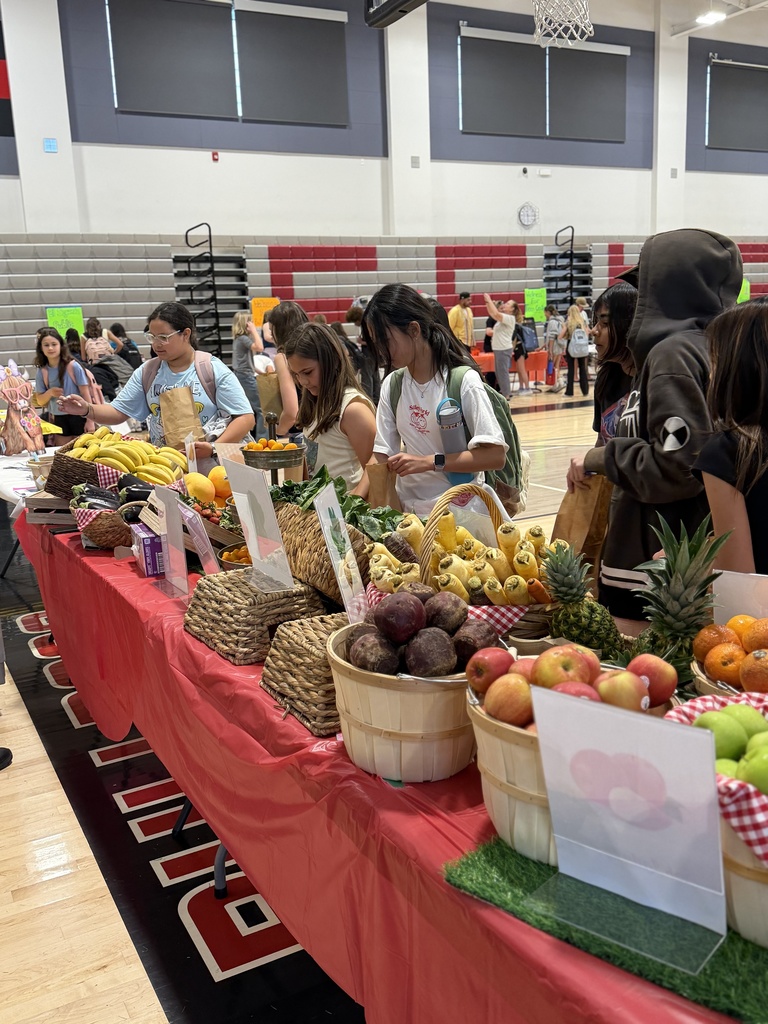 Students line up at a produce table in a gym, choosing fruits and vegetables while holding brown paper bags.
