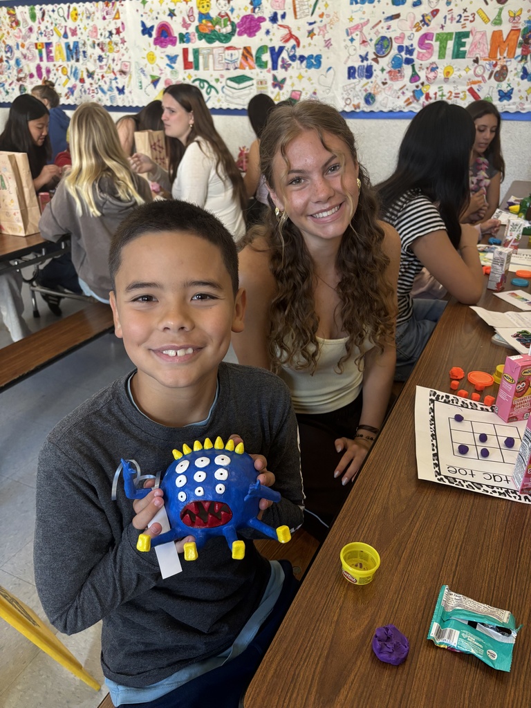 Child holding a colorful blue plush toy with multiple eyes and yellow spikes in a busy classroom setting.