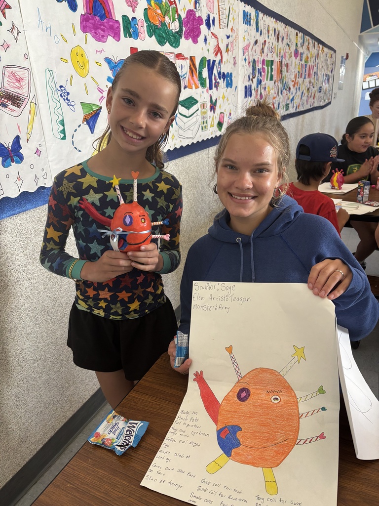 Two children display a colorful drawing of an orange creature with multiple arms and legs in a classroom decorated with student artwork.