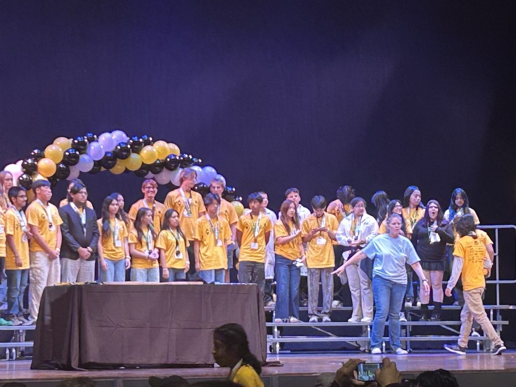 Group of young people wearing yellow shirts standing on stage under black, white, and gold balloon arch during event.