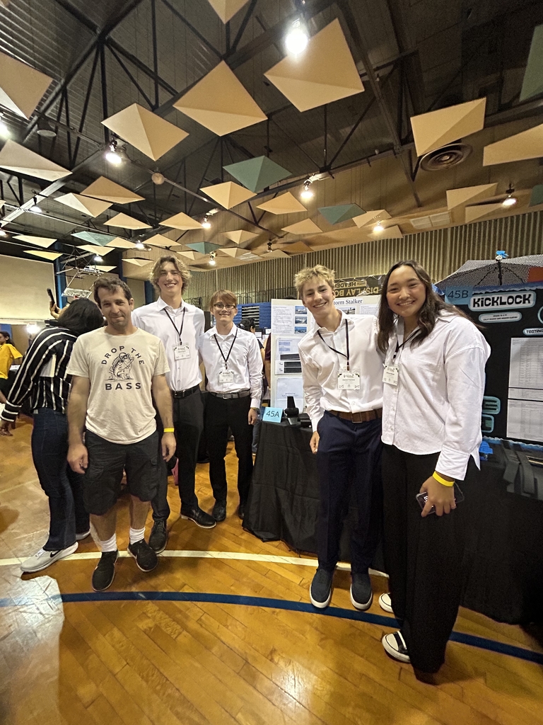 Group of five young people standing in a gymnasium with a science project display behind them at a school event.