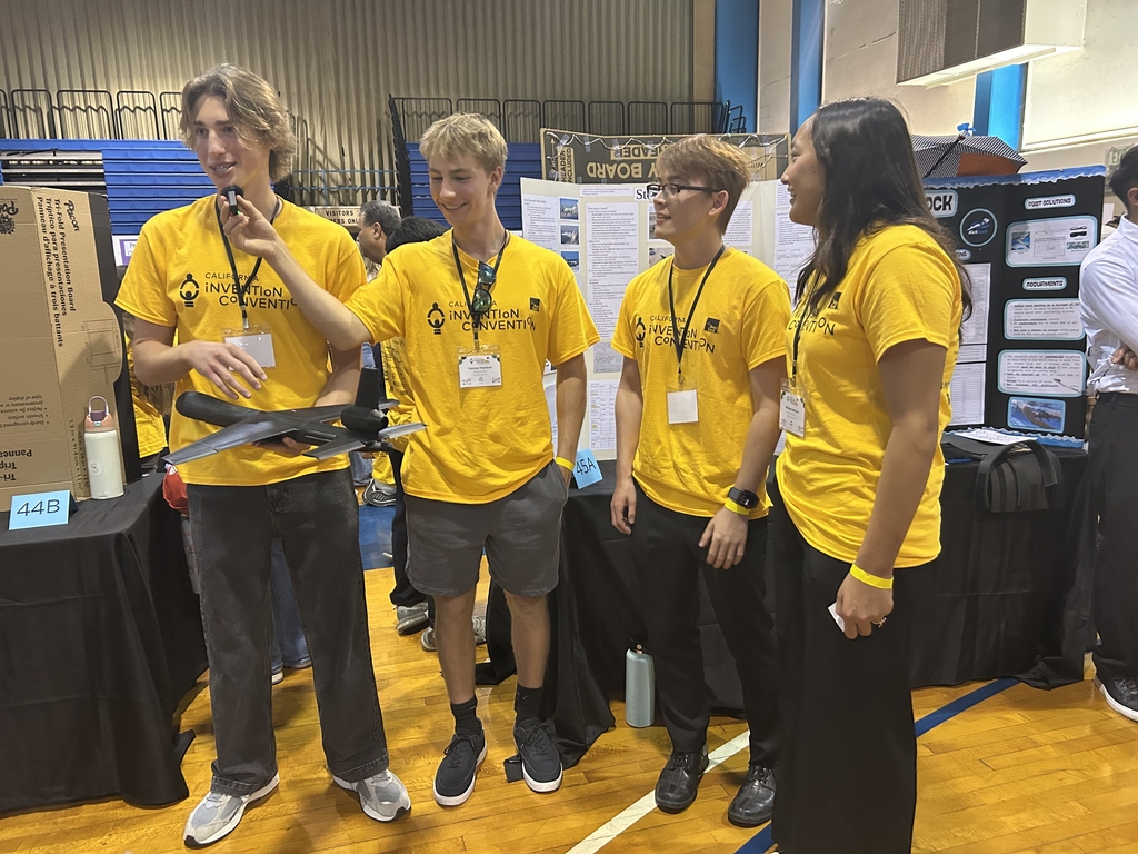 Four students wearing matching yellow Innovation Convention t-shirts stand together in a gymnasium presenting their project at a science fair.