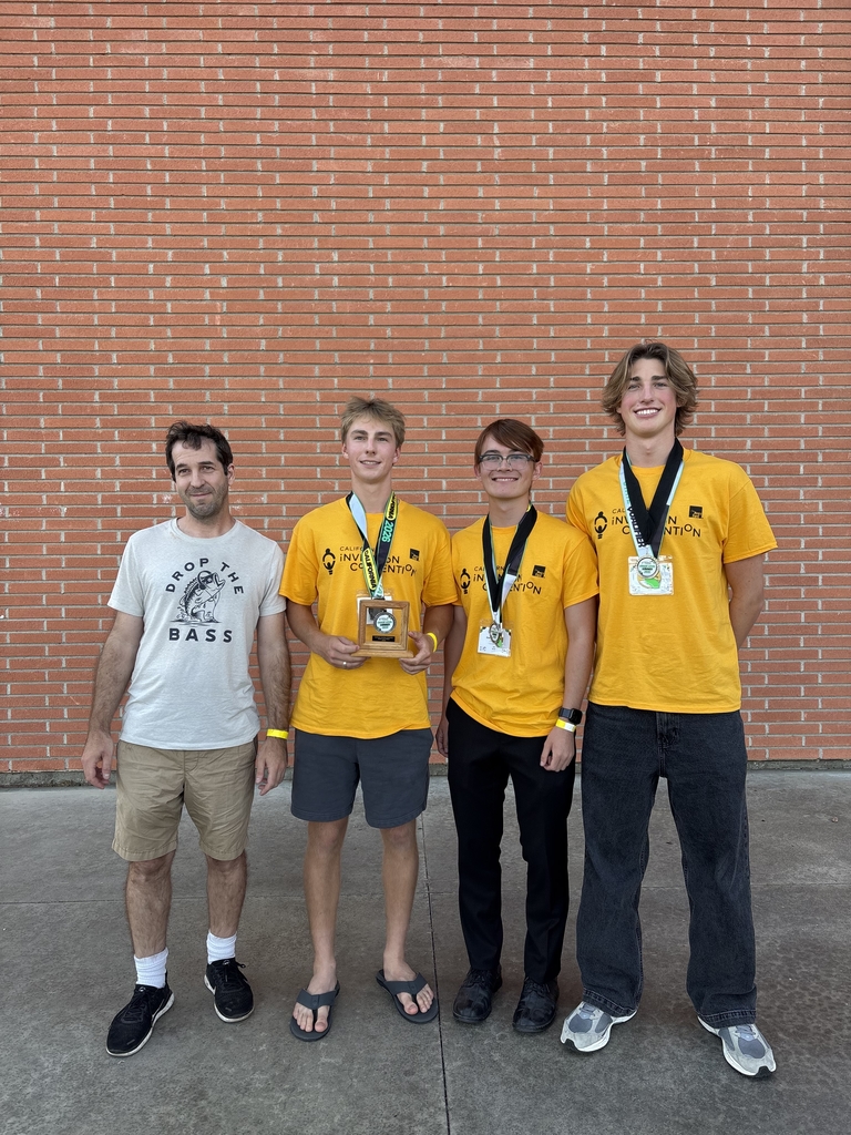 Four young men standing in front of a brick wall, three wearing yellow shirts with medals and one holding a trophy, celebrating a competition win.