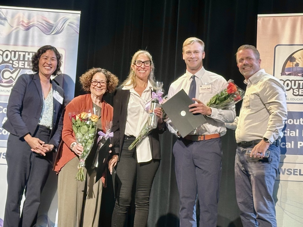 Five adults stand on stage smiling and holding certificates and bouquets, celebrating an award or recognition event.
