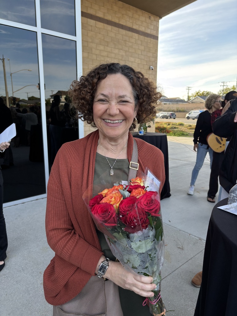 Smiling woman holds a bouquet of red and orange roses outdoors near a building during a celebration or recognition gathering.