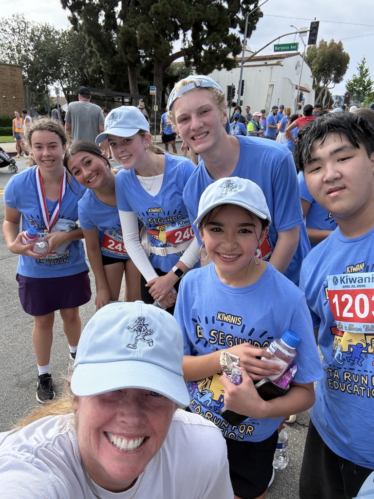Group of runners wearing matching blue Kiwanis race shirts and white caps posing together outdoors after a race event.