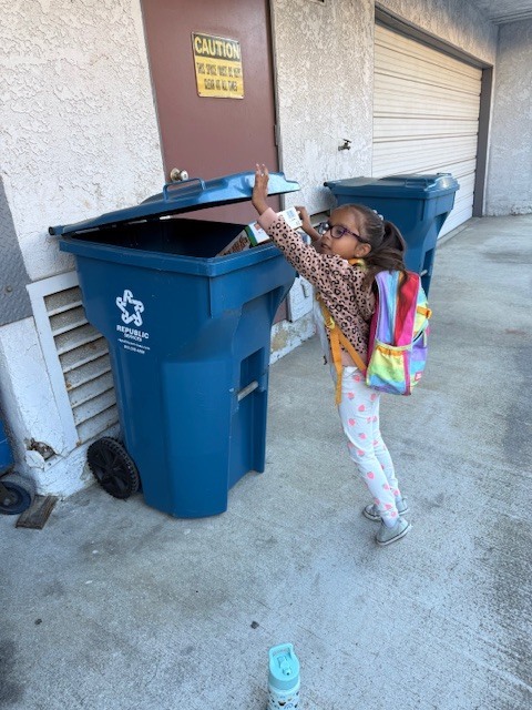 Young girl with backpack reaches into a large outdoor recycling bin, placing an item inside near a building wall.