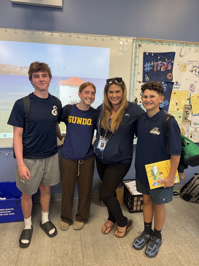Teacher stands with three students in a classroom, smiling in front of a projected coastal image on the board.