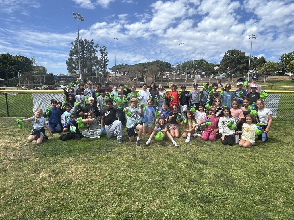 Large group of students pose on a grassy field holding green bags and gloves, likely after a cleanup activity.