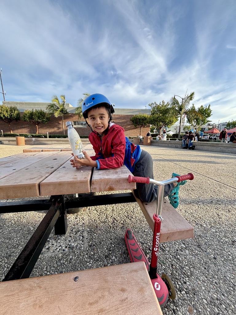 Young child in helmet and red hoodie leans on a picnic table at a park, holding a water bottle beside a scooter.
