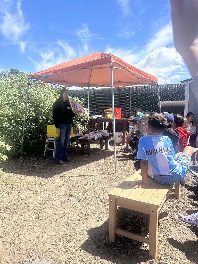 Woman speaking to a group of children seated outdoors under a bright orange canopy on a sunny day.