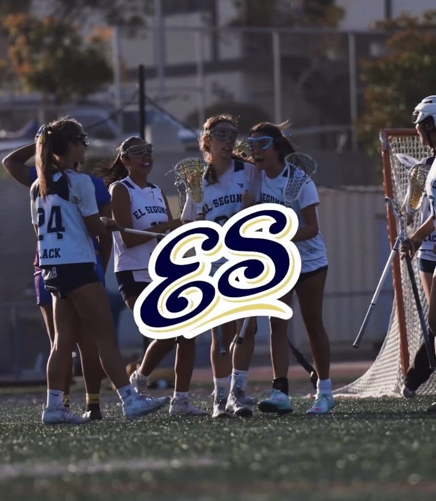 Young female lacrosse players in white El Segundo jerseys gather on the field near the goal during a game.
