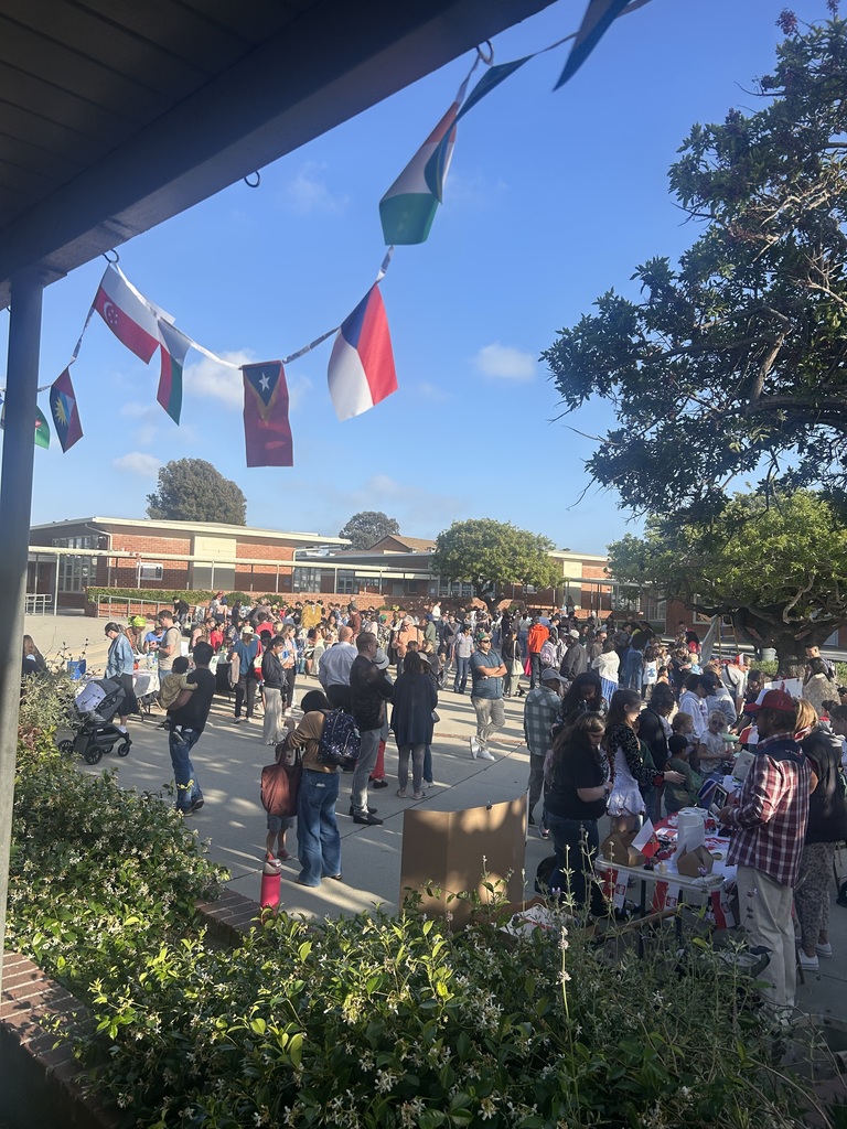 Crowd of people gathered outdoors near a school building under a clear blue sky with international flags hanging above.