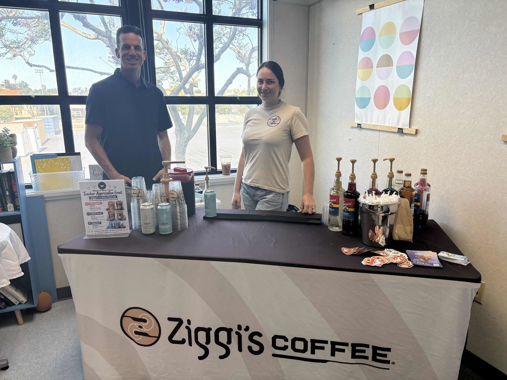 Two baristas stand behind a Ziggi’s Coffee setup with syrups, drinks, and event signage near large windows.