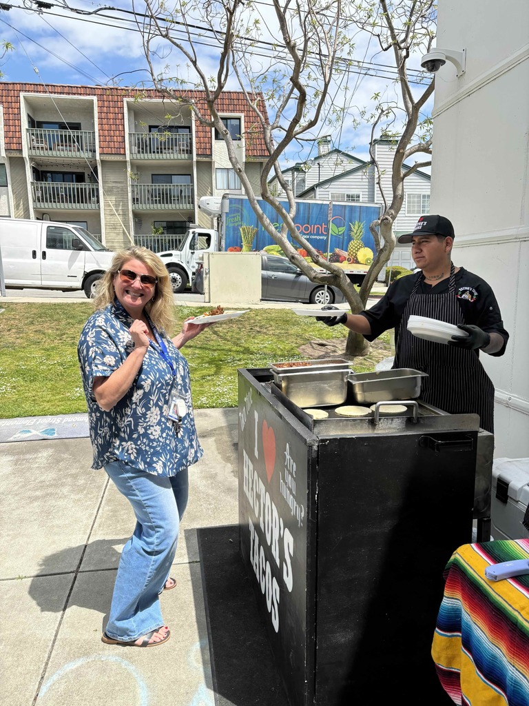 Staff member smiles while receiving tacos from an outdoor food station during a sunny appreciation event.